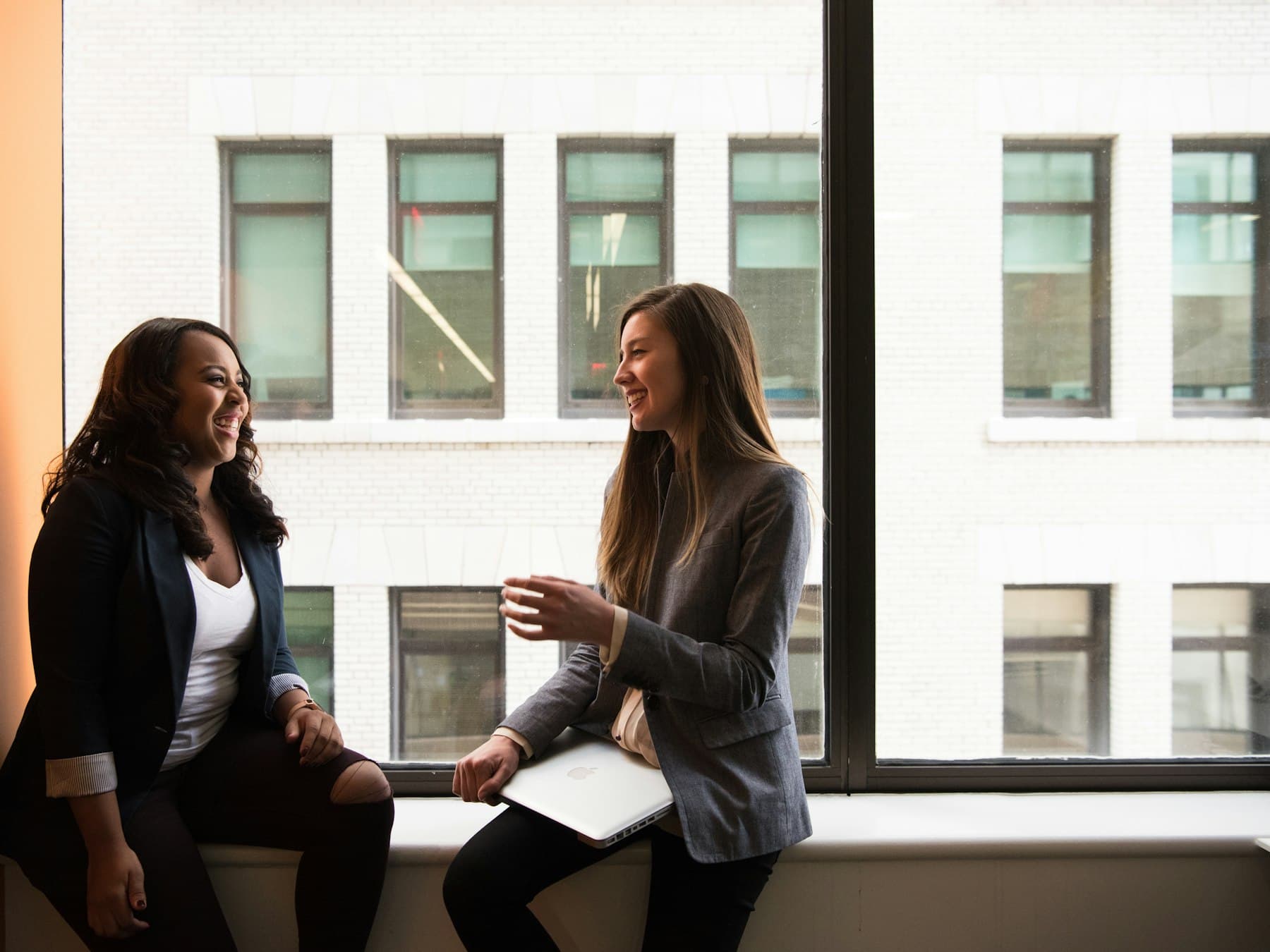 Two colleagues laughing together while chatting on a sunny window ledge