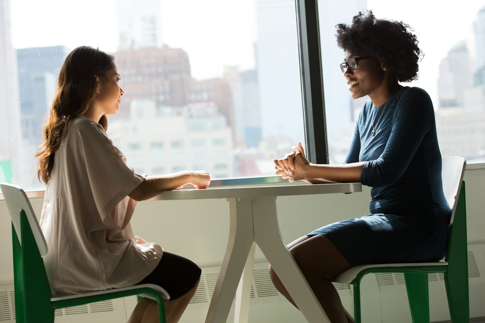 Two women talking across a small table by a sunny window