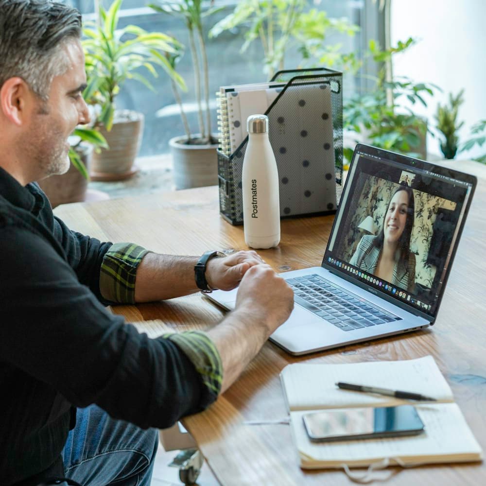 A man on a video call from a laptop at a sunny table surrounded by plants
