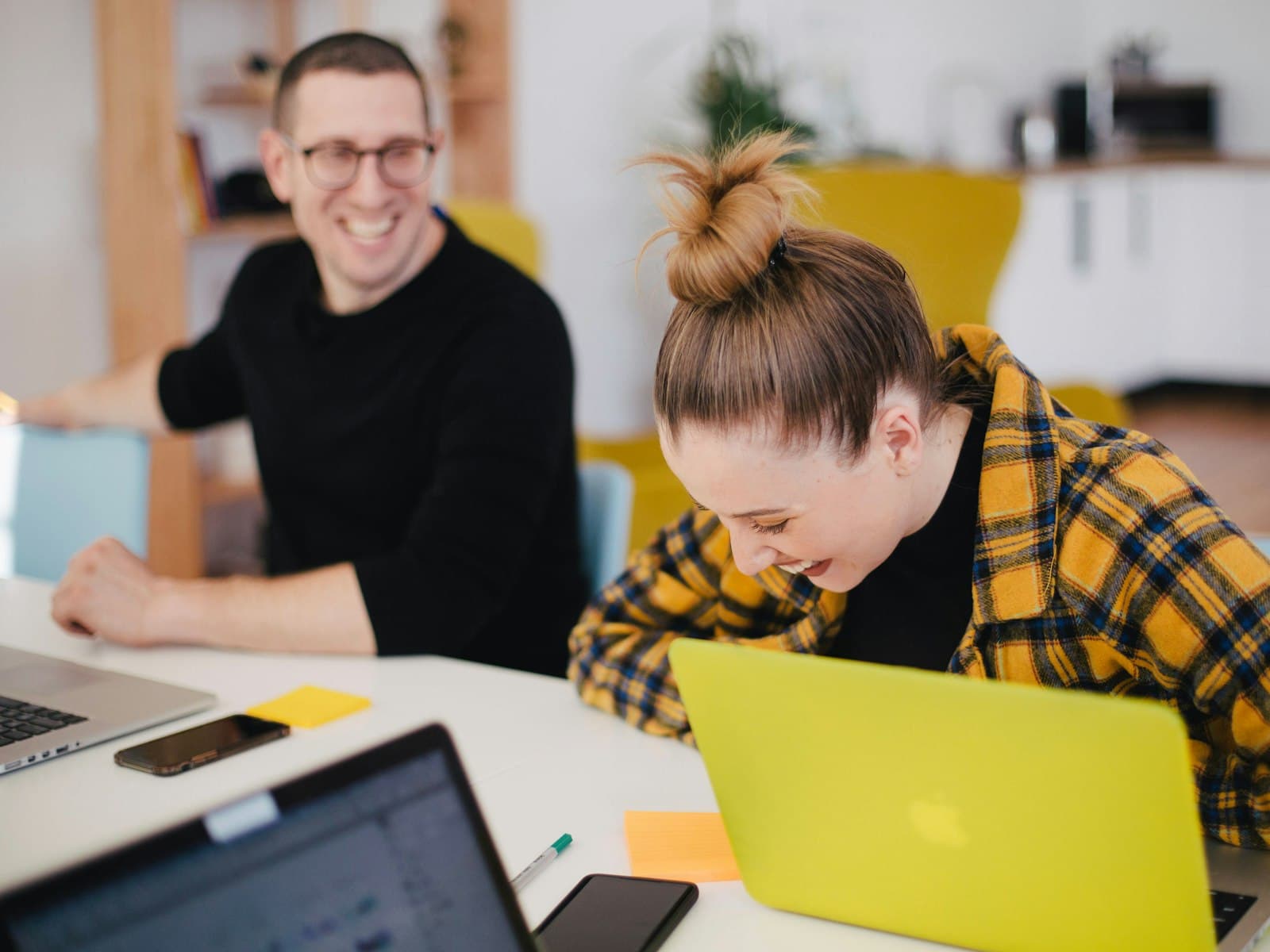 Two colleagues laughing together while working on laptops in a bright office